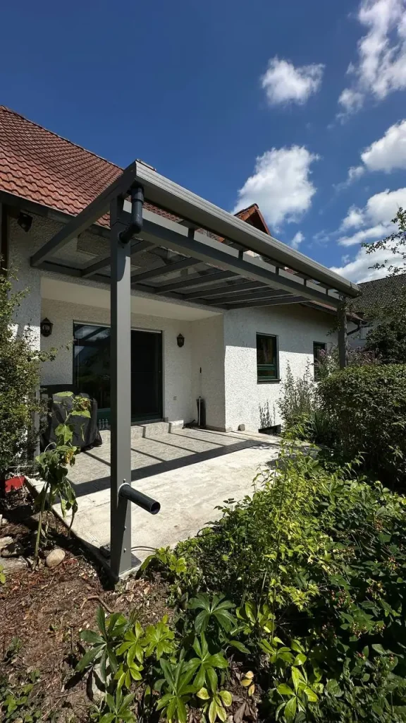 Terrassenüberdachung aus Aluminium mit Glasdach an einer Hausecke mit Blumenbeet