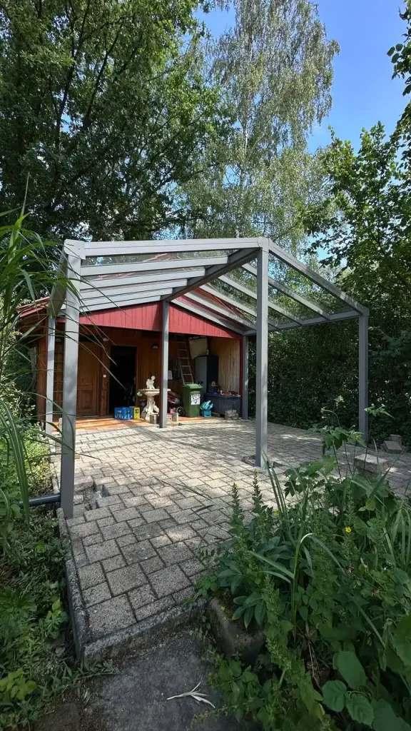 Alu-Terrassenüberdachung mit Glasdach zwischen Bäumen neben einem Holzhaus